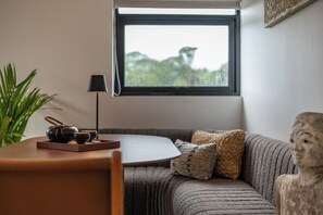 A room with a round table, a black lamp, a sofa with patterned cushions, and a window with a view of greenery.