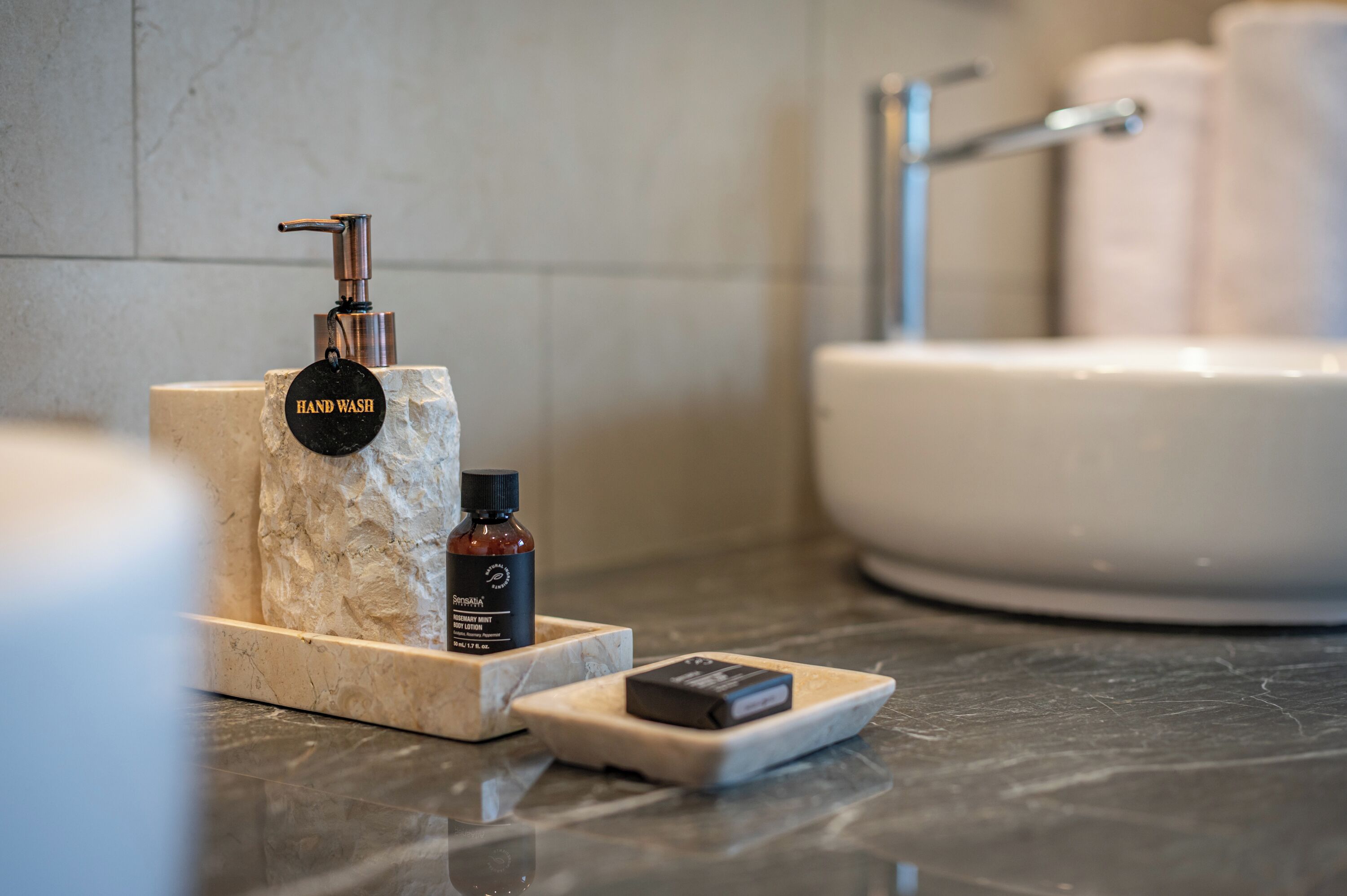Un tocador de baño con un lavamanos, una botella de loción corporal de romero y menta, y un jabonera.