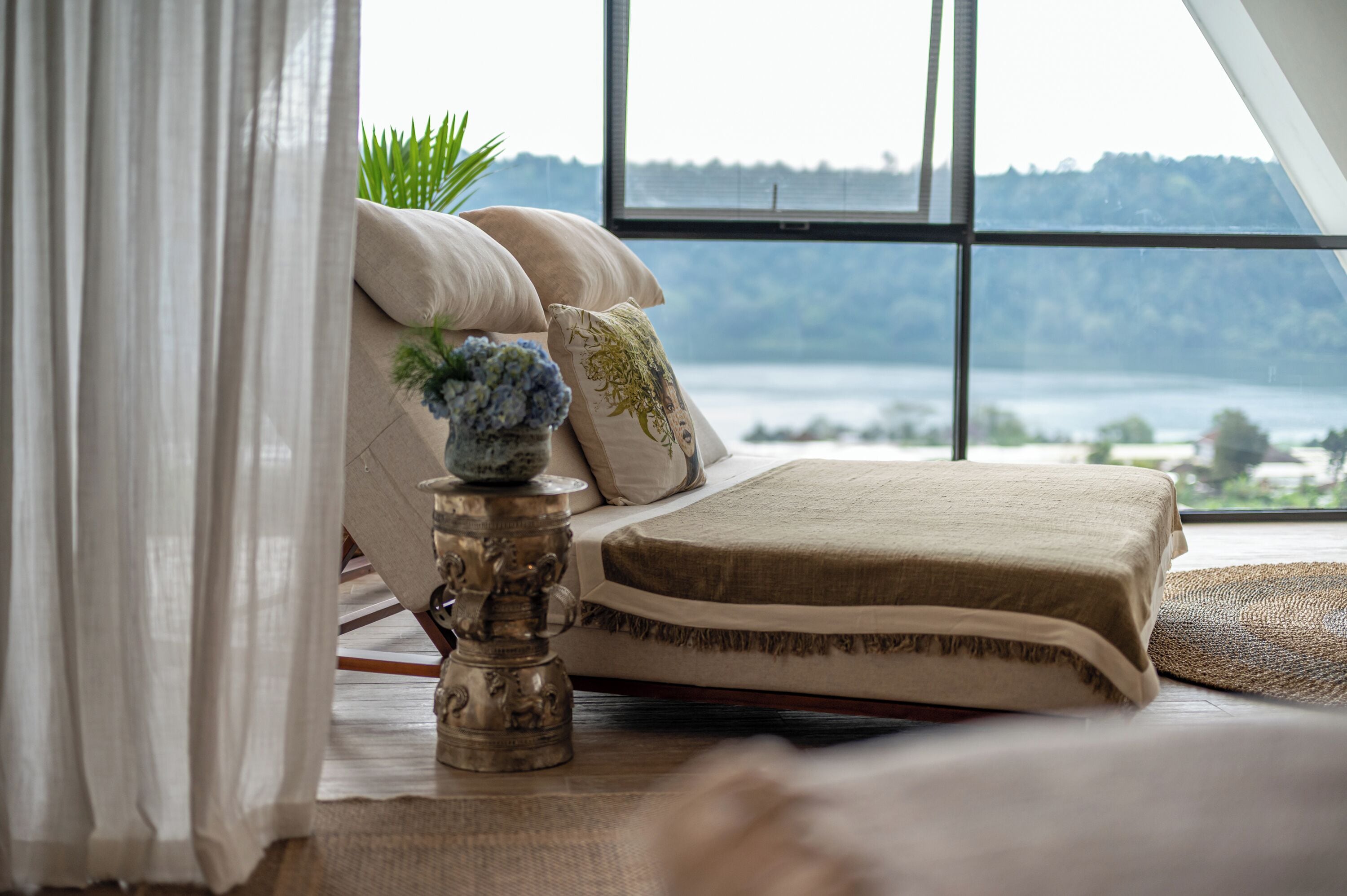 A room with a bed, a window with a view of a body of water and hills, and a decorative lamp on the floor.