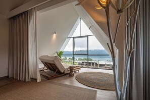 A room with a large window, a lounge chair, a small table, and a view of a body of water.