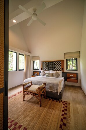 Un dormitorio con una cama grande, una silla de madera, un ventilador de techo y una ventana con vistas a zonas verdes.