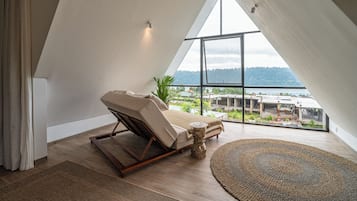 A modern bedroom with a sloped ceiling, a reclining chair, a round woven rug, and a view of a landscape through large windows.