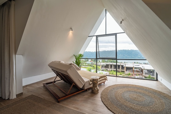 A modern bedroom with a sloped ceiling, a reclining chair, a round woven rug, and a view of a landscape through large windows.