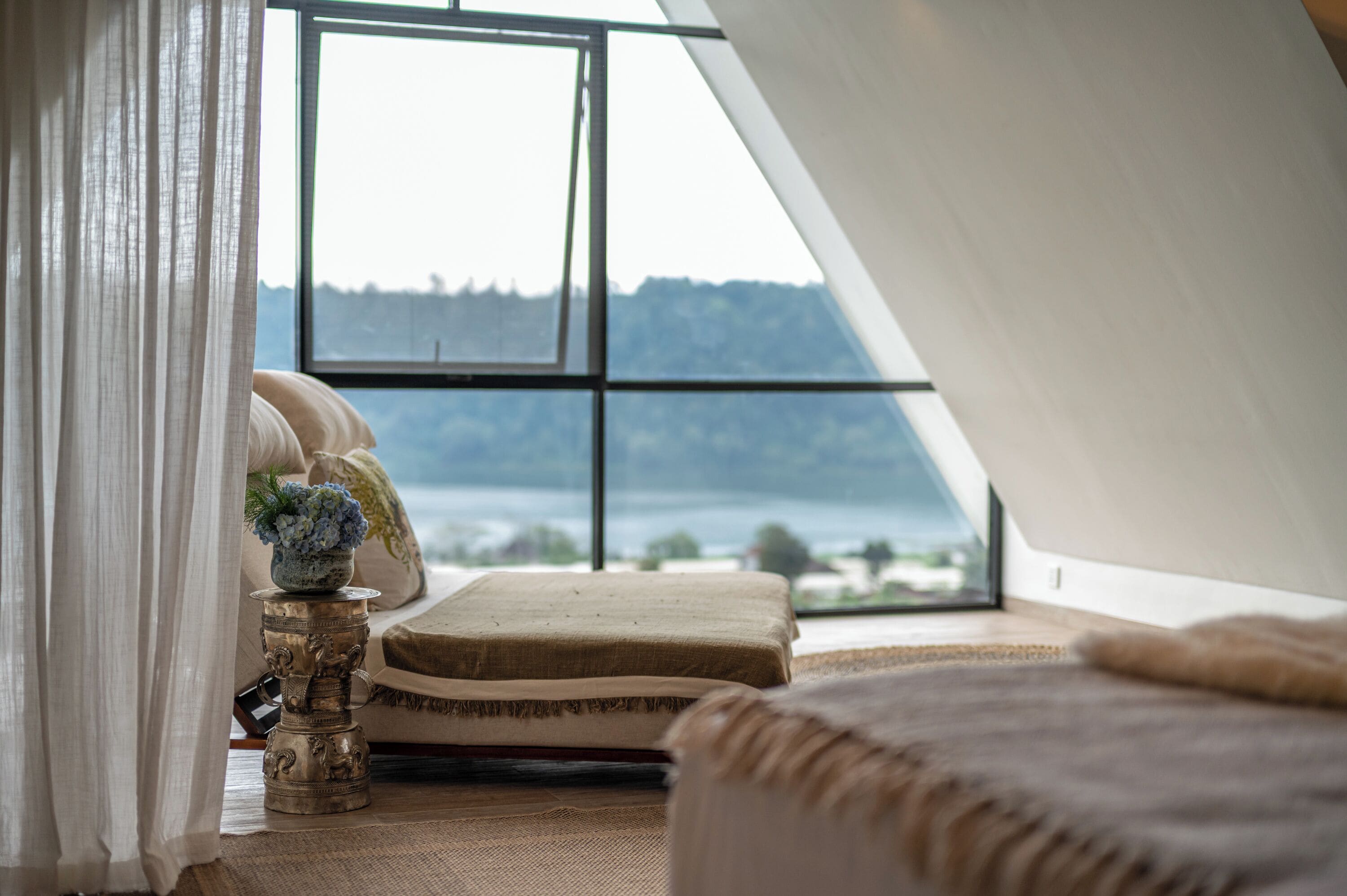 A bedroom with a large window, a bed, a decorative metal stand, and a view of a body of water and hills.