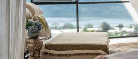 A room with a sloped ceiling, a bed with a cushion, a decorative metal pot, and a view of a landscape through a large window.