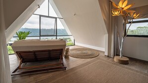 A room with a high ceiling, a large window, a wooden bench, a woven rug, and a tall floor lamp.