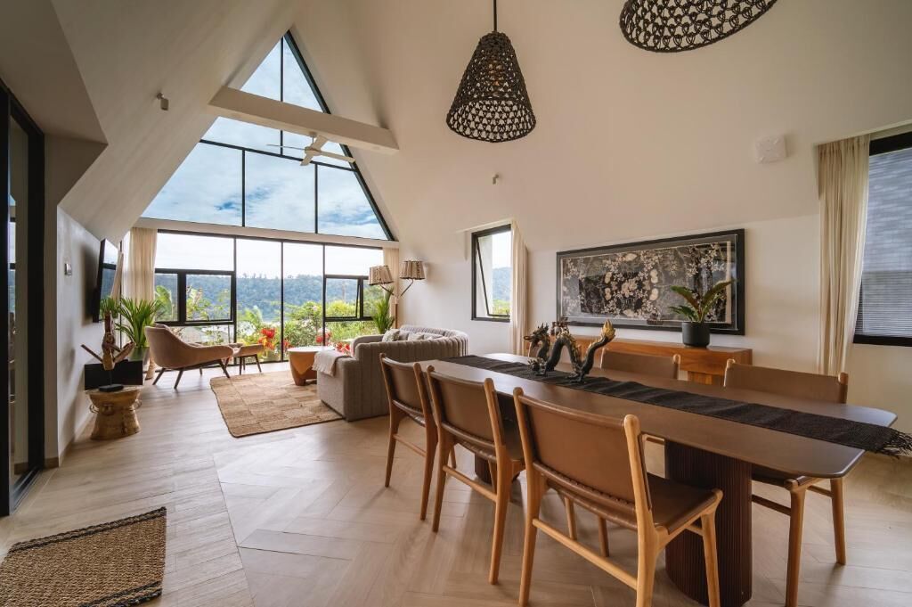 A spacious room with a high ceiling, a dining table with chairs, a sofa, and a large window offering a view of the outdoors.