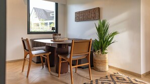 A dining area with a round table, four wooden chairs, a potted plant, and a view of a house through a window.