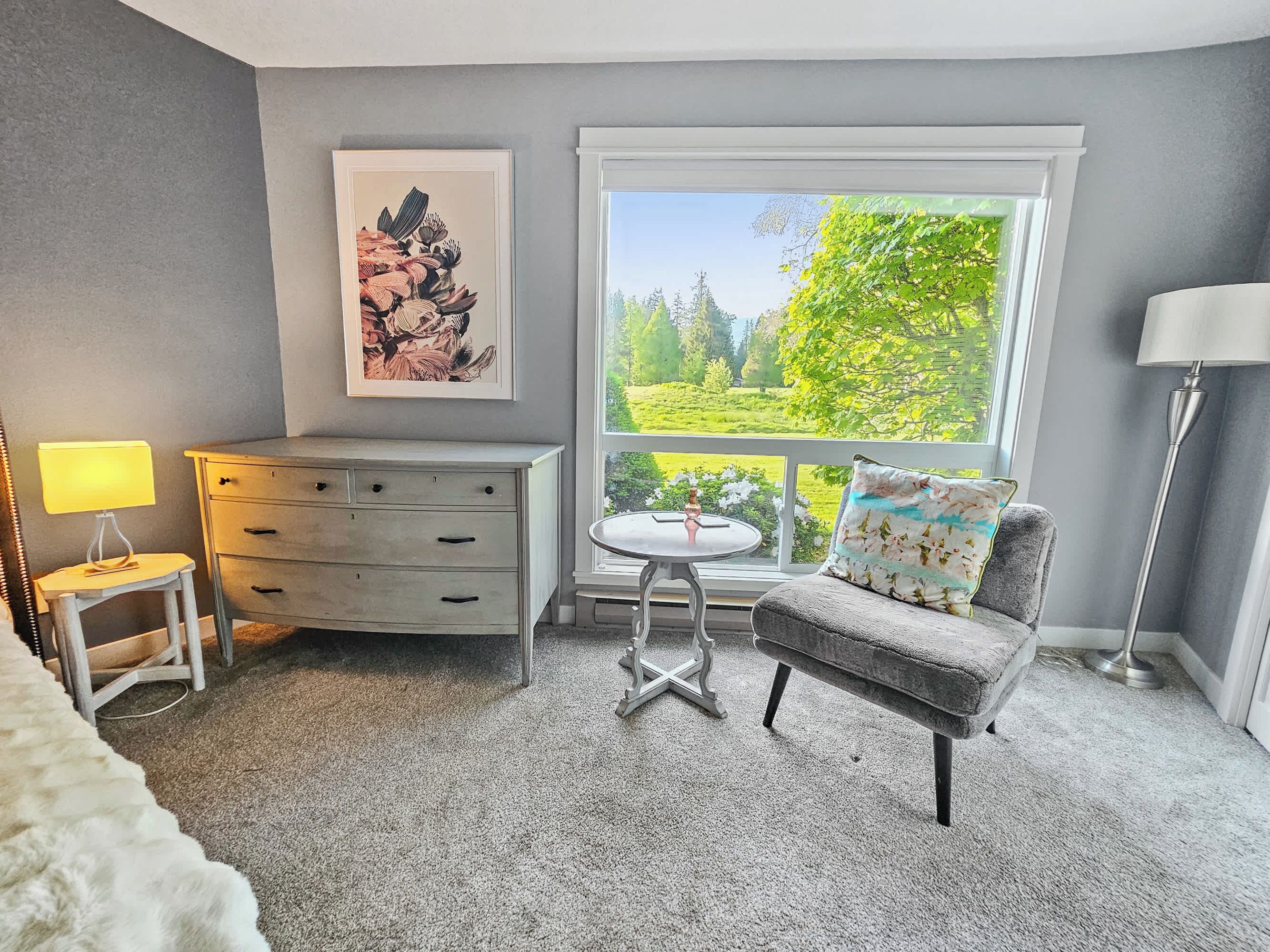 Une chambre avec une grande fenêtre, une commode, une chaise et une lampe.