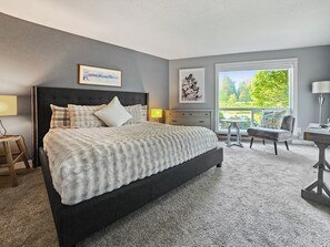 A bedroom with a large bed, a nightstand, a chair, and a desk. There is a window with a view of trees. - Mt Hood Condo Near Skiing & Golf Hawaiian-Inspired (Mount Hood Village)