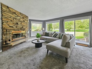 Ein Wohnzimmer mit Kamin aus Stein, einer Eckcouch und Blick auf eine grüne Landschaft durch große Fenster.