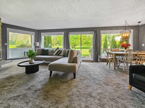 A spacious living room with a sectional sofa, a dining area with wooden chairs, and large windows offering a view of a garden. - Mt Hood Condo Near Skiing & Golf Hawaiian-Inspired (Mount Hood Village)