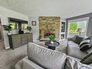 A living room with a stone fireplace, grey sofas, a coffee table, and a view of greenery outside. - Mt Hood Condo Near Skiing & Golf Hawaiian-Inspired (Mount Hood Village)