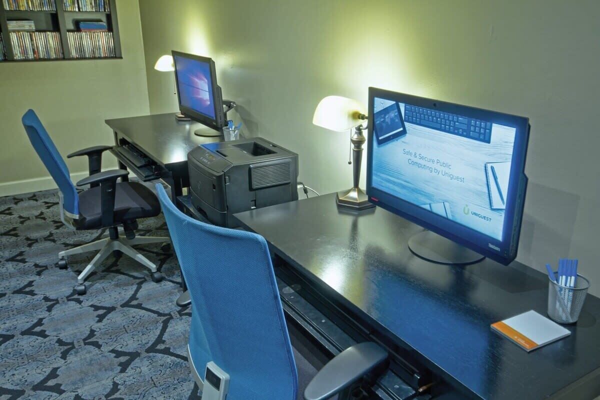 A hotel room with a desk, two monitors, a printer, a lamp, and a chair.