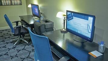 A hotel room with a desk, two monitors, a printer, a lamp, and a chair.