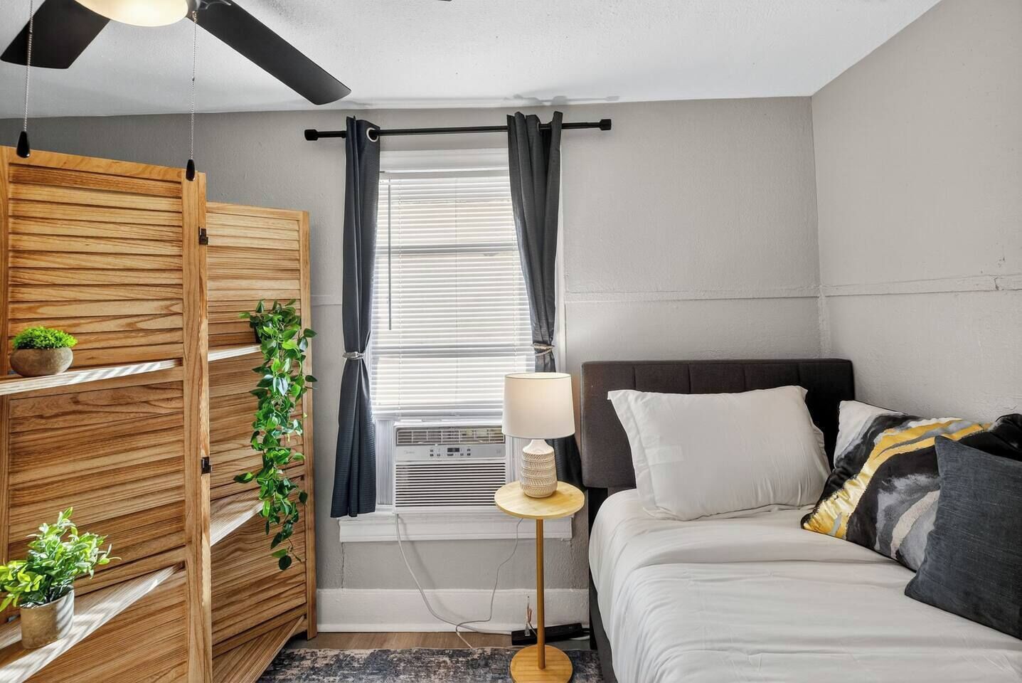 Una camera da letto con un letto, comodino, lampada, finestra con tende e un armadio in legno con piante.