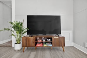 A flat-screen TV on a wooden TV stand with storage compartments, a potted plant, and a white wall. - Minimalist Escape | 12 mins to Downtown Houston (Houston)