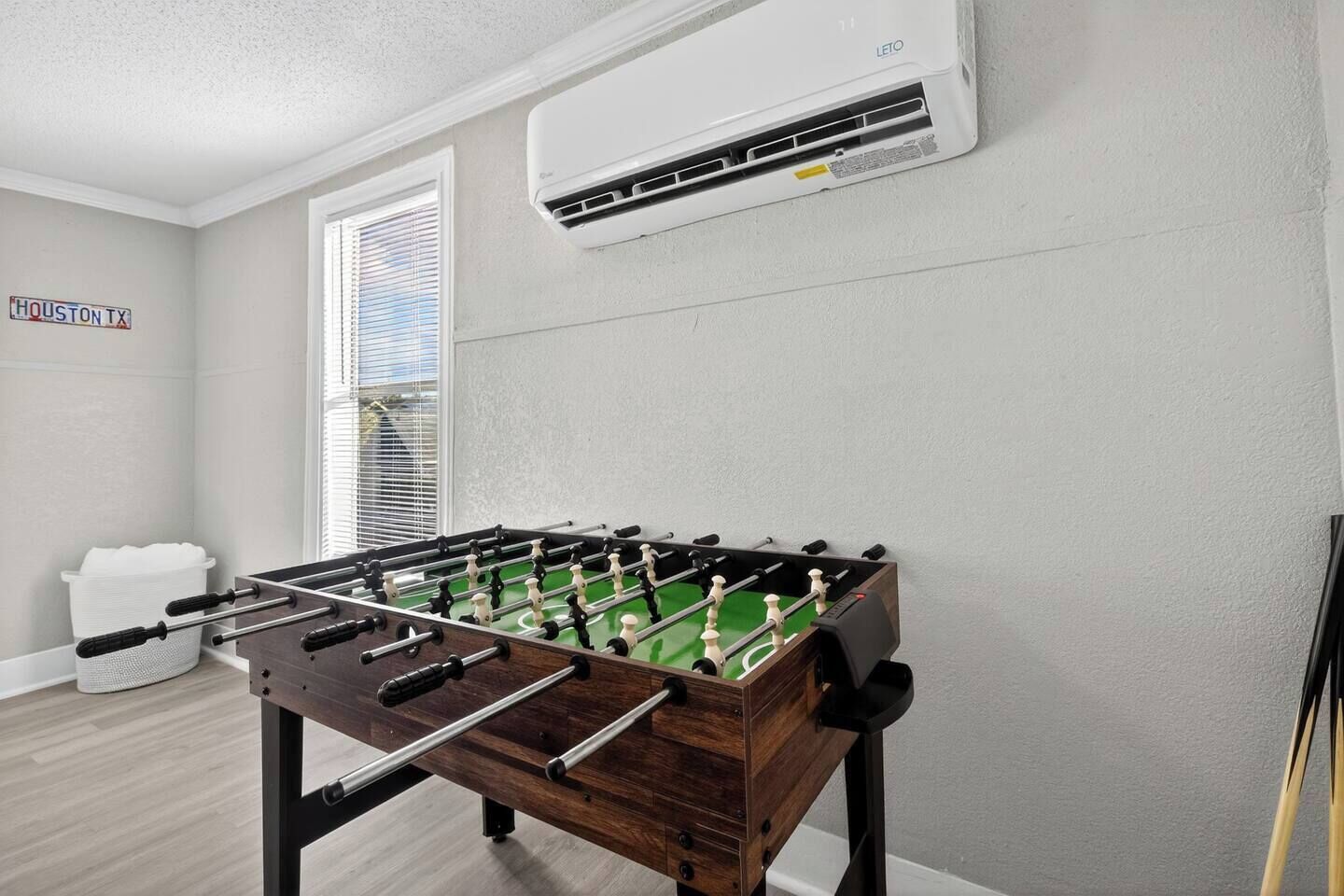 Een kamer met een tafelvoetbaltafel, een raam met zonwering, een luchtverkoeler aan de muur en een bord waarop 'HOUSTON TX' staat.