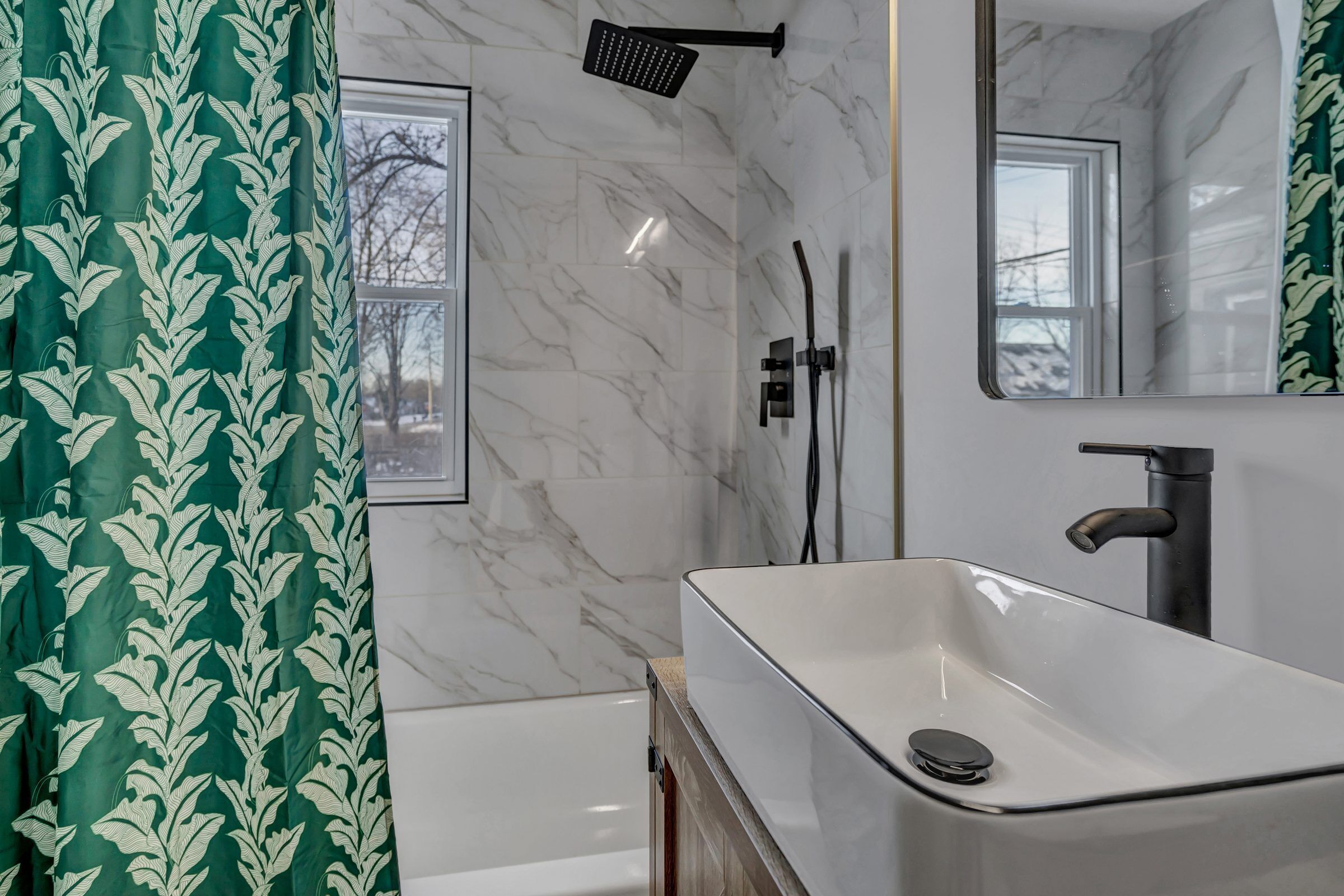 Un baño con lavamanos blanco, un espejo, cabezal de ducha y ventana.