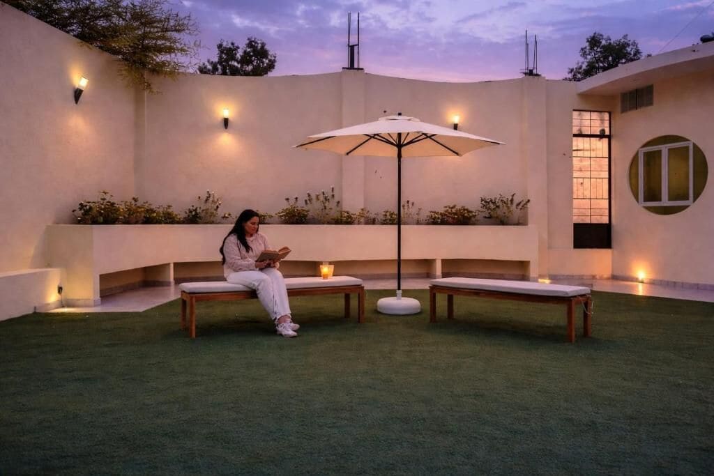 Una persona sentada en un banco en un espacio exterior bien iluminado, con una mesa y una sombrilla.