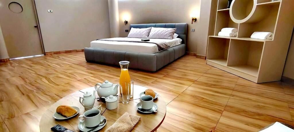 Une chambre d’hôtel avec un lit, une table pour le petit-déjeuner avec du thé, du café et du jus de fruits, et une étagère avec des serviettes.