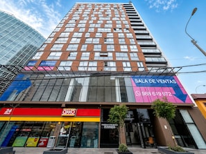 A modern building with a glass facade, a visible storefront with a red awning, and a sign for 'Sala de Ventas' with a phone number. - Depa moderno en Miraflores Gym y parking (Lima)