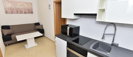 Una cocina compacta con horno integrado, un lavamanos y un microondas. Incluye un refrigerador pequeño, una encimera blanca y una puerta de madera que da a otra habitación.