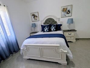 Ein Schlafzimmer mit einem weißen Bett, zwei Nachttischen mit Lampen, einem blauen Vorhang und zwei an der Wand hängenden Bildern.