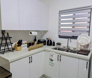 Eine Küche mit weißen Schränken, einem Spülbecken, einem Fenster mit Jalousien, einem Toaster, einem Wasserkocher und einem Schild mit der Aufschrift 'Life is short, lick the bowl'.