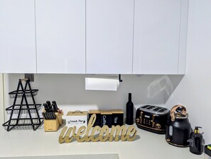 Eine Küchenarbeitsplatte mit einem 'Welcome'-Schild, einem Messerblock, einem Korb, einer mit 'Snack Bar' beschrifteten Kiste, einer Flasche, einem Toaster und einem Wasserkocher.