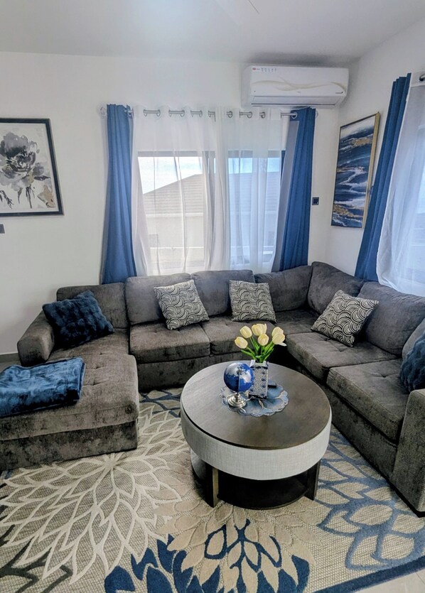 A living room with a grey sectional sofa, patterned cushions, a round coffee table with a globe and vase of flowers, and a patterned rug.