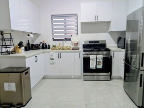 Eine moderne Küche mit weißen Schränken, einem Edelstahl-Kühlschrank, einem Einbauofen und einem Fenster mit Jalousien.