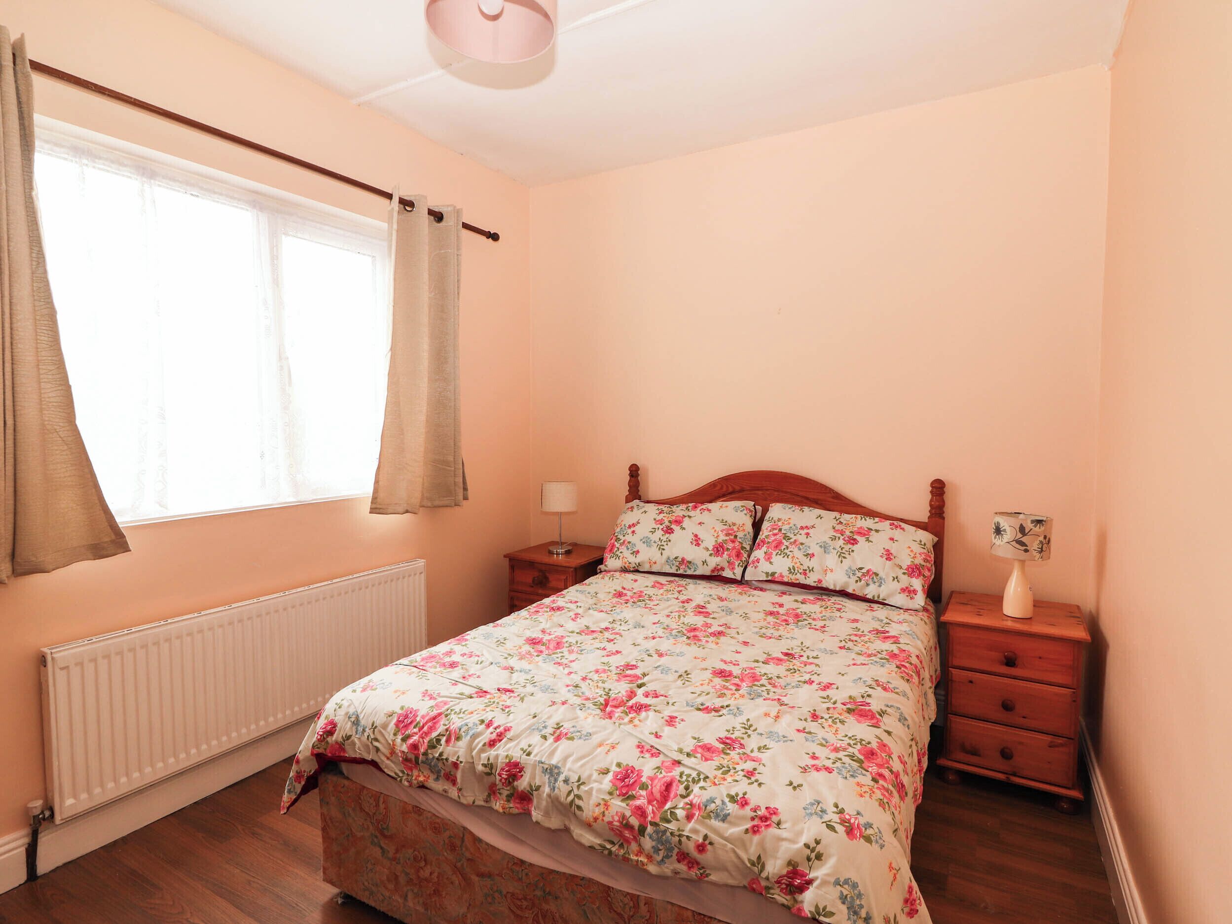 Une chambre à coucher avec un lit en bois, une table de chevet, une commode, une fenêtre avec des rideaux et un radiateur.