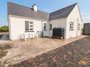 A white two-story house with a grey roof, a small patio with white plastic chairs, and a black water tank. - GOLF CLUB VIEW, pet friendly, with open fire in Doonbeg, County Clare (County Clare)