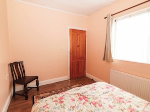 A bedroom with a wooden door, a chair, a floral bedspread, and a window with curtains. - GOLF CLUB VIEW, pet friendly, with open fire in Doonbeg, County Clare (County Clare)