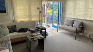 A living room with a sofa, a chair, a coffee table, and a sliding glass door leading to an outdoor area. - Iconic comfortable idyllic bach/cottage, in secluded Enclosure Bay , (Waiheke Island)