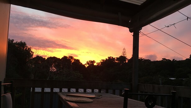 A wooden deck with a table and chairs, overlooking a sunset with trees in the background. - Iconic comfortable idyllic bach/cottage, in secluded Enclosure Bay , (Waiheke Island)