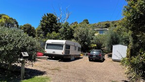 A gravel driveway leading to a white RV and a black car parked in a residential area with trees and a shed. - Iconic comfortable idyllic bach/cottage, in secluded Enclosure Bay , (Waiheke Island)