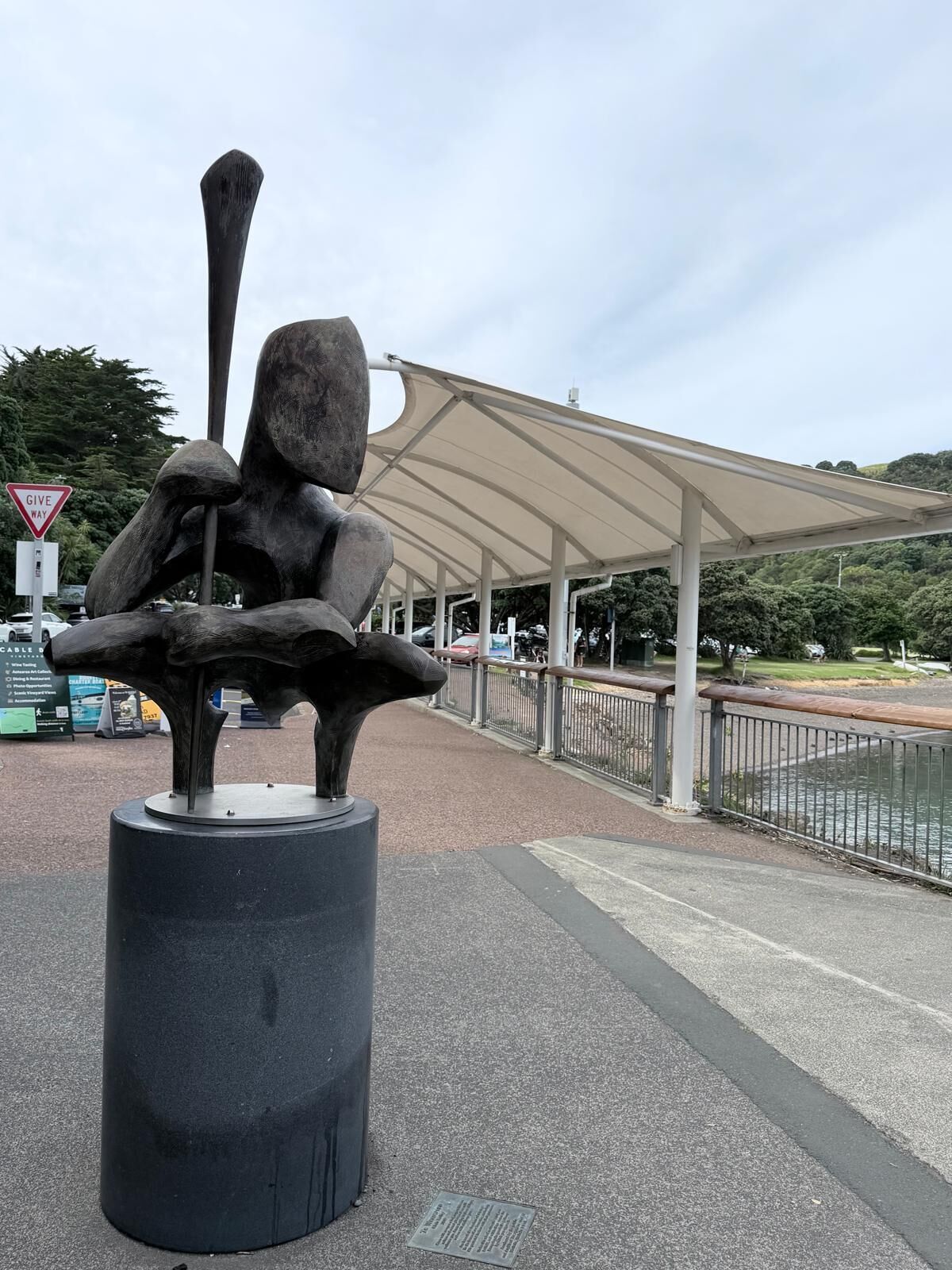 En skulptur som föreställer en figur med utsträckta armar, placerad på en piedestal i ett offentligt område med en överbyggd gångväg och en vattenyta i bakgrunden.