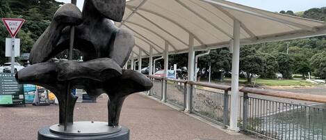 Escultura de una figura con los brazos extendidos sobre un pedestal, en un espacio público con un paseo techado y un cuerpo de agua al fondo.