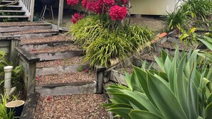 A house with a wooden balcony, steps leading to the entrance, and a variety of plants. - Iconic comfortable idyllic bach/cottage, in secluded Enclosure Bay , (Waiheke Island)