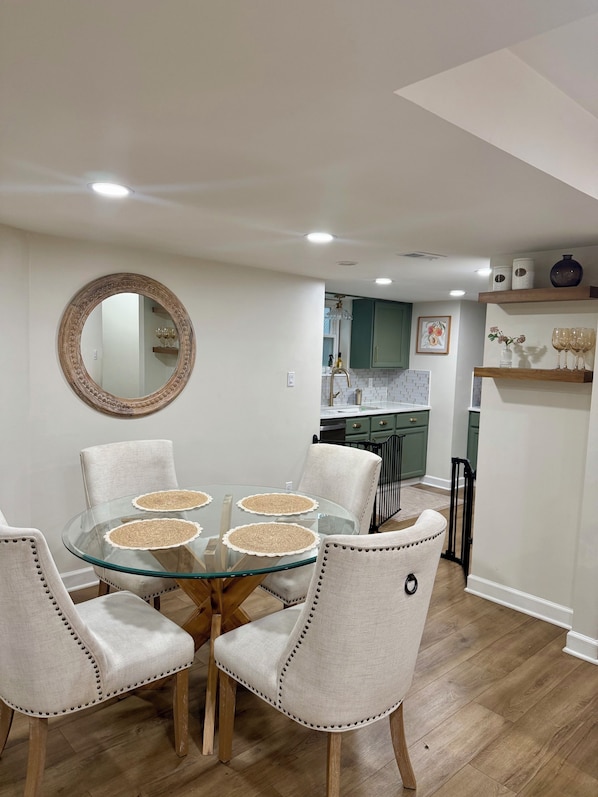 A dining area with a glass table surrounded by upholstered chairs, a round mirror on the wall, and a kitchen in the background.