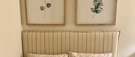 Un lit recouvert d’une courtepointe beige, deux oreillers à motifs floraux et, au-dessus, deux reproductions botaniques encadrées accrochées au mur.