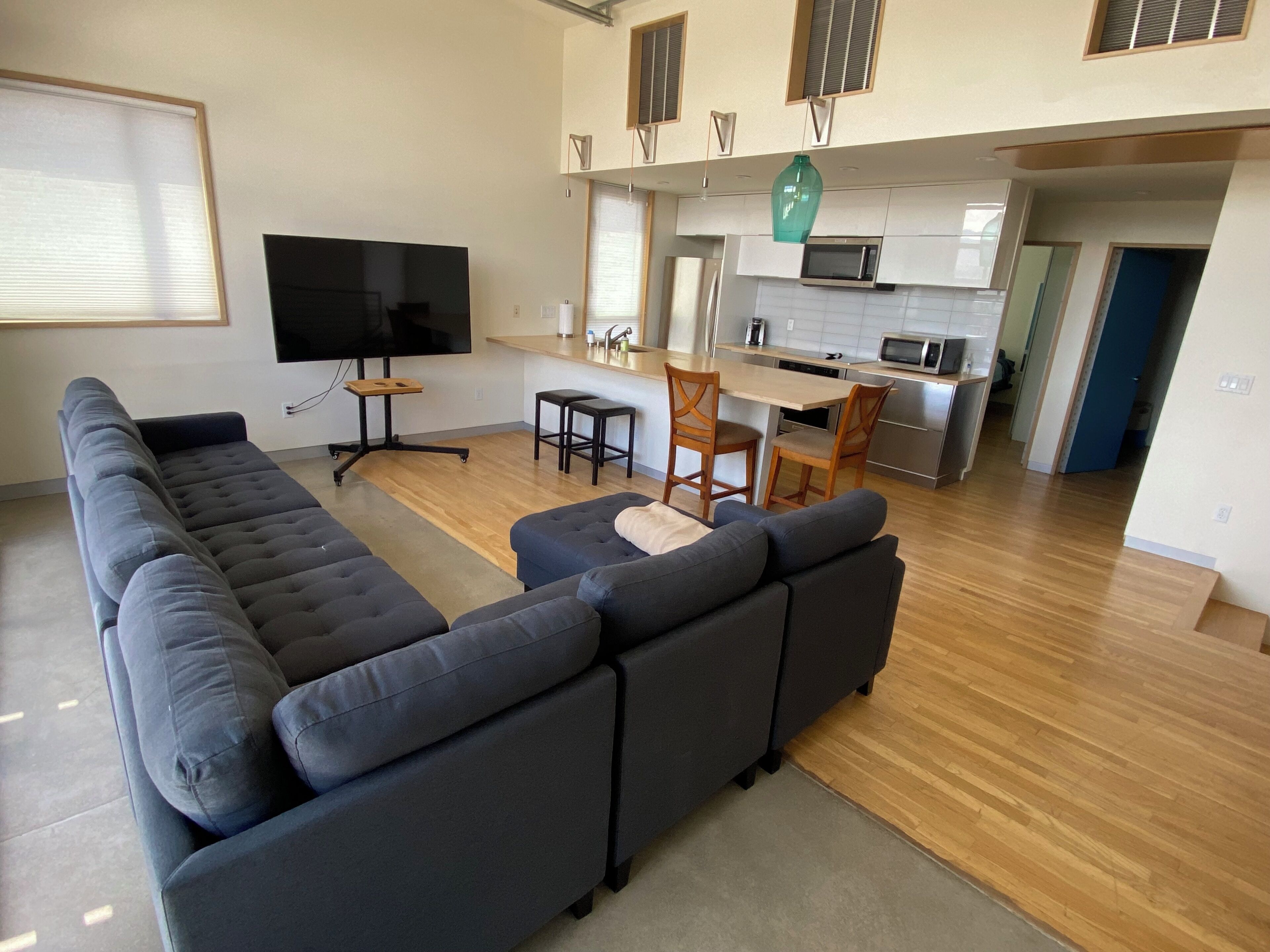 A modern living area with a large sectional sofa, a flat-screen TV, and a kitchen with a microwave and sink.