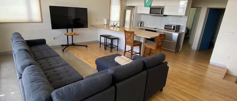 A modern living area with a large sectional sofa, a flat-screen TV, and a kitchen with a microwave and sink.