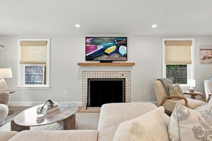 Ein Wohnzimmer mit Kamin, einem darüber angebrachten Fernseher und zwei mit Jalousien versehenen Fenstern.