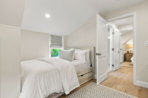 Quarto com cama, mesa de cabeceira, janela com persianas e porta que dá para outro cômodo.