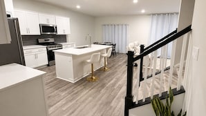 A modern kitchen with white cabinetry, a central island with bar stools, a dining area with a table and chairs, and a staircase with a railing. - Spacious 5BR Upper Home Near DC, National Harbor & Joint Base Andrews (Waldorf)