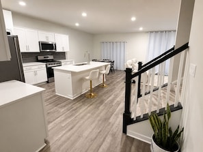 A modern kitchen with white cabinetry, a central island with bar stools, a dining area with a table and chairs, and a staircase with a railing.