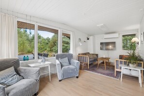 A modern living room with a sofa, armchair, and a TV. There are windows with curtains, a wooden floor, and a small table. - Cozy holiday home in Hune near Blokhus (Hune)